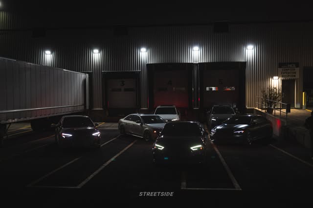 A row of Audis parked nose-out in front of numbered loading-dock doors at night, headlights on.